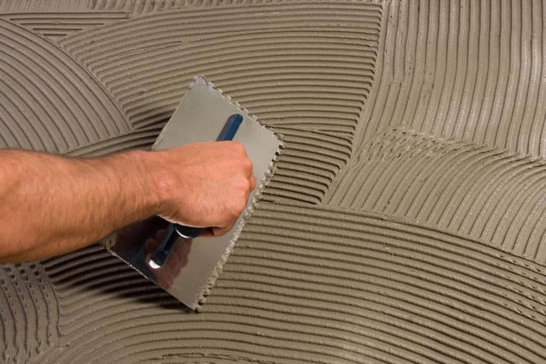 Man's hand combing a layer of mortar, using a trowel's notched side.